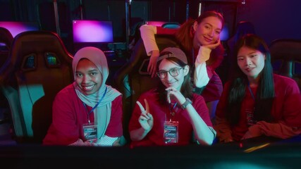 Medium portrait of happy multiethnic female gamer team in red t-shirts, with tournament participant badges, posing together at cyberclub, looking at camera, smiling with victory gesture