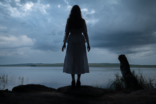 Mysterious woman in white dress standing by a dark lake under stormy sky