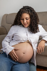 A joyful pregnant woman with curly hair smiles proudly as she gently holds her baby bump, dressed casually in jeans and a white blouse, celebrating motherhood and new beginnings.