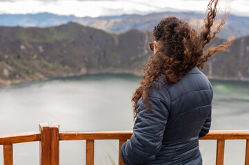 A woman with curly brown hair stands at a viewpoint overlooking the breathtaking Quilotoa Lake. Her peaceful stance captures a quiet moment of reflection and natural beauty.
