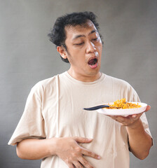 Portrait of handsome asian man eat instant noodle on grey background. He's feel surprise and...