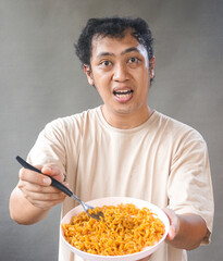 Portrait of handsome asian man eat instant noodle on grey background. He's feel surprise and...