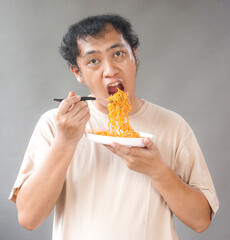 Portrait of handsome asian man eat instant noodle on grey background. He's feel surprise and...