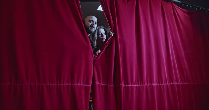 Two young actors peek through red stage curtains, excited and ready for their performance. They step onto the stage, smiling and eager to captivate the audience.