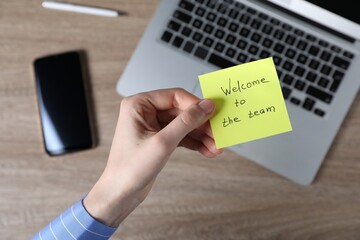Woman holding paper note with words Welcome To The Team at wooden table, top view
