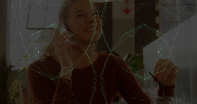 Talking woman in sweater holding phone, lifting glass with straw at cafe table with network overlay
