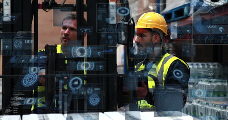 Two men wearing vests, helmet operating forklift controls in warehouse with shelving and pallets
