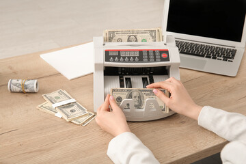 Woman putting dollar banknotes into cash counting machine at table in office, closeup