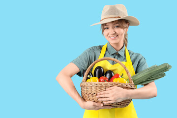 Happy young female farmer with wicker basket full of different ripe vegetables on blue background