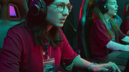 Medium tilt-up shot of geeky young Caucasian girl wearing thick eyeglasses, baseball cap, participant badge competing in multiplayer video game with female team, excitedly talking to teammates