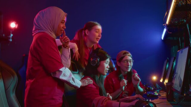 Medium side shot of young Asian girl sitting at computer in cyberclub, playing shooter match online, multiethnic friends standing around, watching. Ideal for international esport championship 