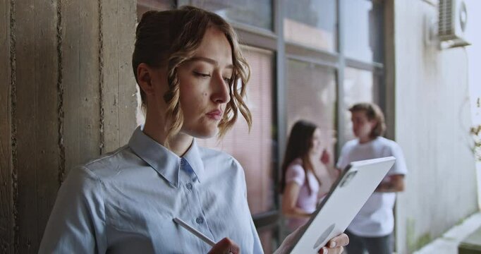 A focused young student utilizes a digital tablet and stylus for her education, demonstrating modern learning methods on a university campus with peers in the background.