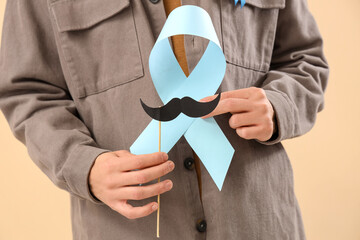 Young man with blue ribbon and paper mustache on beige background, closeup. Prostate cancer...