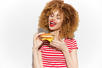 Joyful young woman with curly hair enjoying a delicious burger, wearing a striped shirt, showcasing...