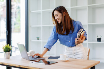 Smiling young asian woman sitting at desk working on laptop taking notes in notebook, happy...
