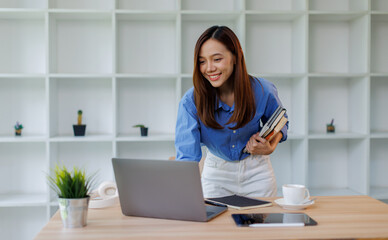Smiling young asian woman sitting at desk working on laptop taking notes in notebook, happy millennial female studying online, watching webinar using computer and writing check list
