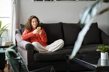 Young woman sitting on couch holding smartphone doing ecommerce shopping, smiling lady looking at...