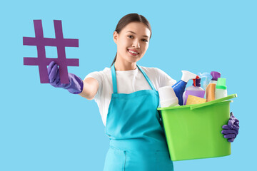 Female janitor with cleaning supplies and hashtag sign on color background