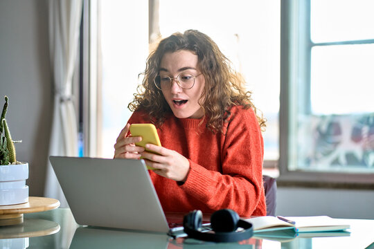 Young surprised woman student feeling amazed looking at smartphone, shocked girl using mobile cell phone reading online message, receiving news or sms offer with positive unexpected news at home.
