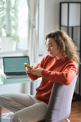 Young pretty woman holding smartphone using cellphone apps modern technology, looking at mobile phone while remote working or e learning from home sitting at table with laptop computer. Vertical.