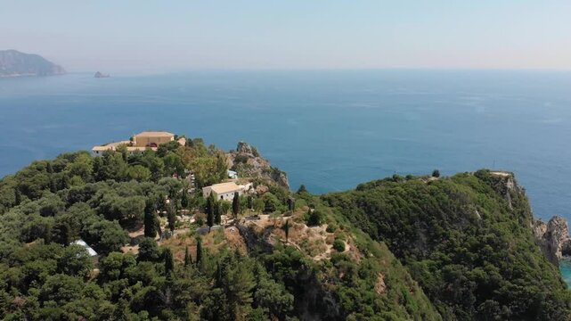Stunning aerial footage showcasing the iconic Paleokastritsa Monastery perched atop a lush green hill, overlooking the crystal clear turquoise waters of the Ionian Sea and the rugged coastline of Corf
