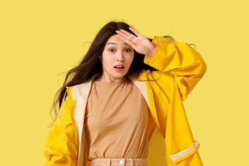 Beautiful Asian woman in raincoat with strong wind on yellow background