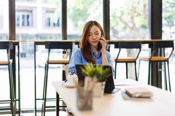 Fototapeta premium Smiling asian woman writing notes while making phone call and using laptop at workplace. 