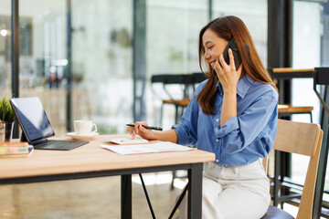 Obraz premium Smiling asian woman writing notes while making phone call and using laptop at workplace. 