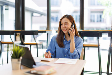 Obraz premium Smiling asian woman writing notes while making phone call and using laptop at workplace. 
