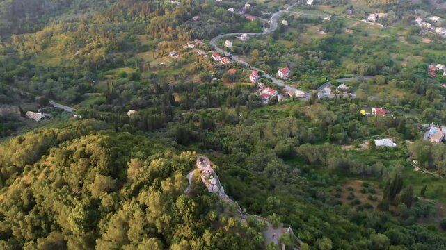 An aerial drone shot captures the panoramic view from Kaiser's Throne (Kaisers Thron) viewpoint in Pelekas village, Corfu Island, Greece, showcasing the lush green hills, scattered residential areas, 