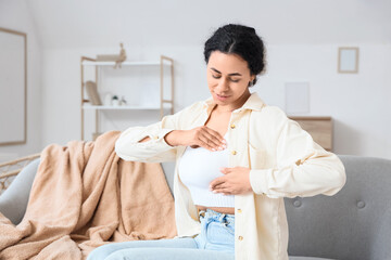 Young African-American woman checking her breast on sofa at home. Cancer awareness concept