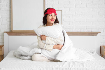 Beautiful young woman with sleep mask, pillow and blanket sitting on bed at home