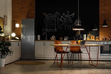 Interior of stylish kitchen with counters, dining table and glowing lamps at night
