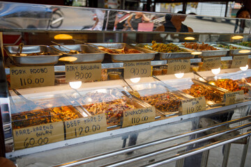 Traditional Filipino cooked meals displayed behind glass at carinderia food stall with handwritten price signs