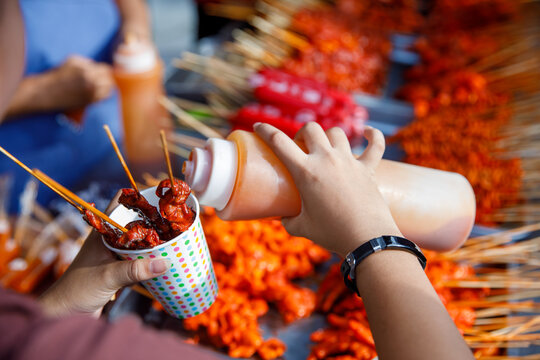Filipino street food skewers with red sauce in paper cup, hand pouring sauce at outdoor market
