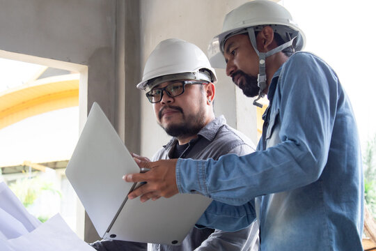 Hispanic engineer man with mustache wearing safety white helmet consult team project manager partnership, Asian foreman standing in construction site building reading blueprint and laptop technology
