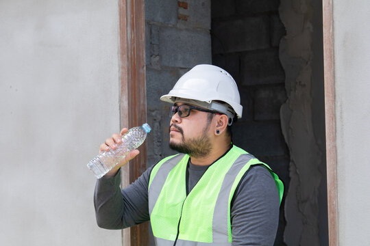 Young Asian engineering worker overweight employee foreman thirsty drinking hydrate water in plastic bottle, foreman worry working on sunny day overheat, heatstroke health care in workplace outdoor