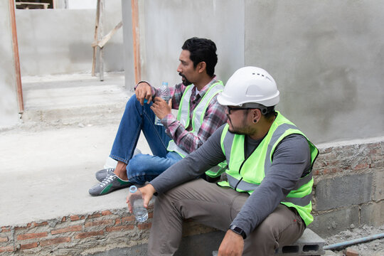 Two Asian builder workers wearing safety reflected vest and helmet tired thirsty hydrated drinking water from plastic bottle together relaxed sitting on construction site on overheated sunny day