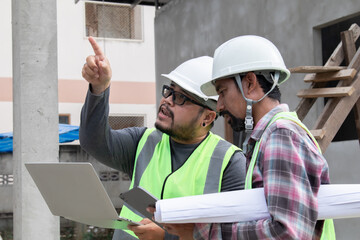 Young overweight engineering foreman reading blueprint paperwork on construction site outdoors with Asian or Indian contractor man partnership project manager using laptop technology solving problem