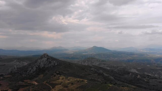 An aerial view captures the dramatic, rugged landscape surrounding Acrocorinth, showcasing the ancient citadel's imposing hill and the vast, undulating terrain of the Peloponnese under a cloudy sky.
