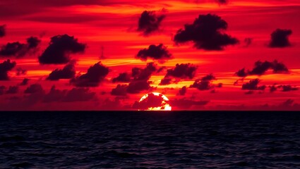 A vibrant red sunset over the ocean with dark clouds scattered across the sky creating a dramatic scene