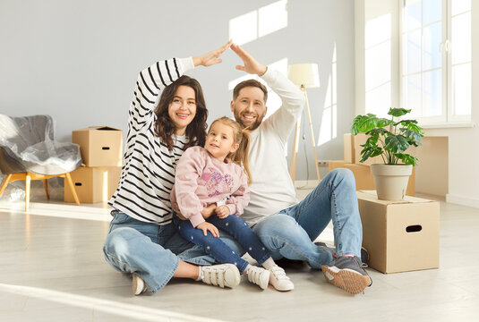 Happy young smiling family with cute daughter making house roof sitting on floor at home among unpacking cardboard boxes on moving day. Insurance, mortgage, real estate and move concept.