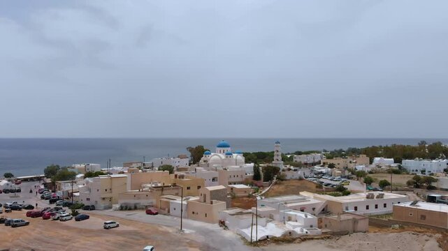 An aerial perspective captures the charming coastal village of Perissa on Santorini, Greece, showcasing its traditional architecture, the iconic blue-domed church, and the expansive black sand beach m