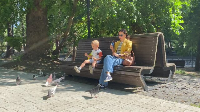 A mother and her young child sit on a bench feeding pigeons. They relax under the shade of large trees in a sunny park in Simferopol, Crimea.