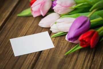 Blank white card is lying next to pink white purple red tulip bouquet on rustic tabletop