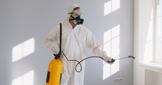 Male exterminator spraying pesticide with sprayer, wearing safety clothes, doing pest control at home, posinng in empty white room. Home disinfection and quarantine during epidemic concept