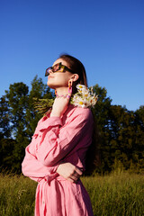 Young woman in pink dress holding flowers, enjoying sunny day outdoors, expressing joy and serenity...