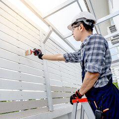 Asian man painter standing on the ladder painting white acrylic color on house wooden wall. Male...