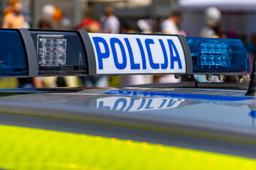 Close-up of a Polish police car light bar with the word "Policja," with blurred figures of people in the background during a public event. © Adam