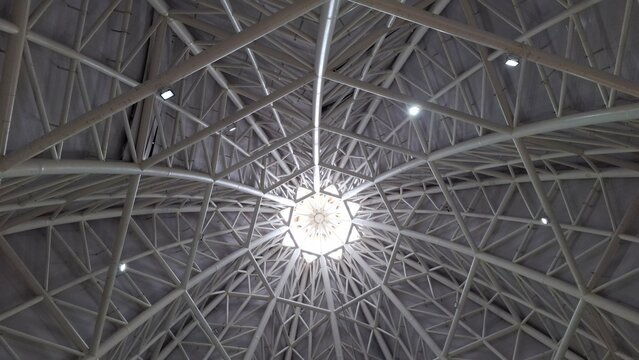 Symmetrical view of a modern architectural ceiling featuring a geometric steel framework and a central skylight. Captured from directly below, showcasing the intricate pattern and industrial design